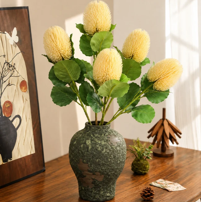 Pale yellow artificial banksia flowers with broad leaves arranged in a green vase